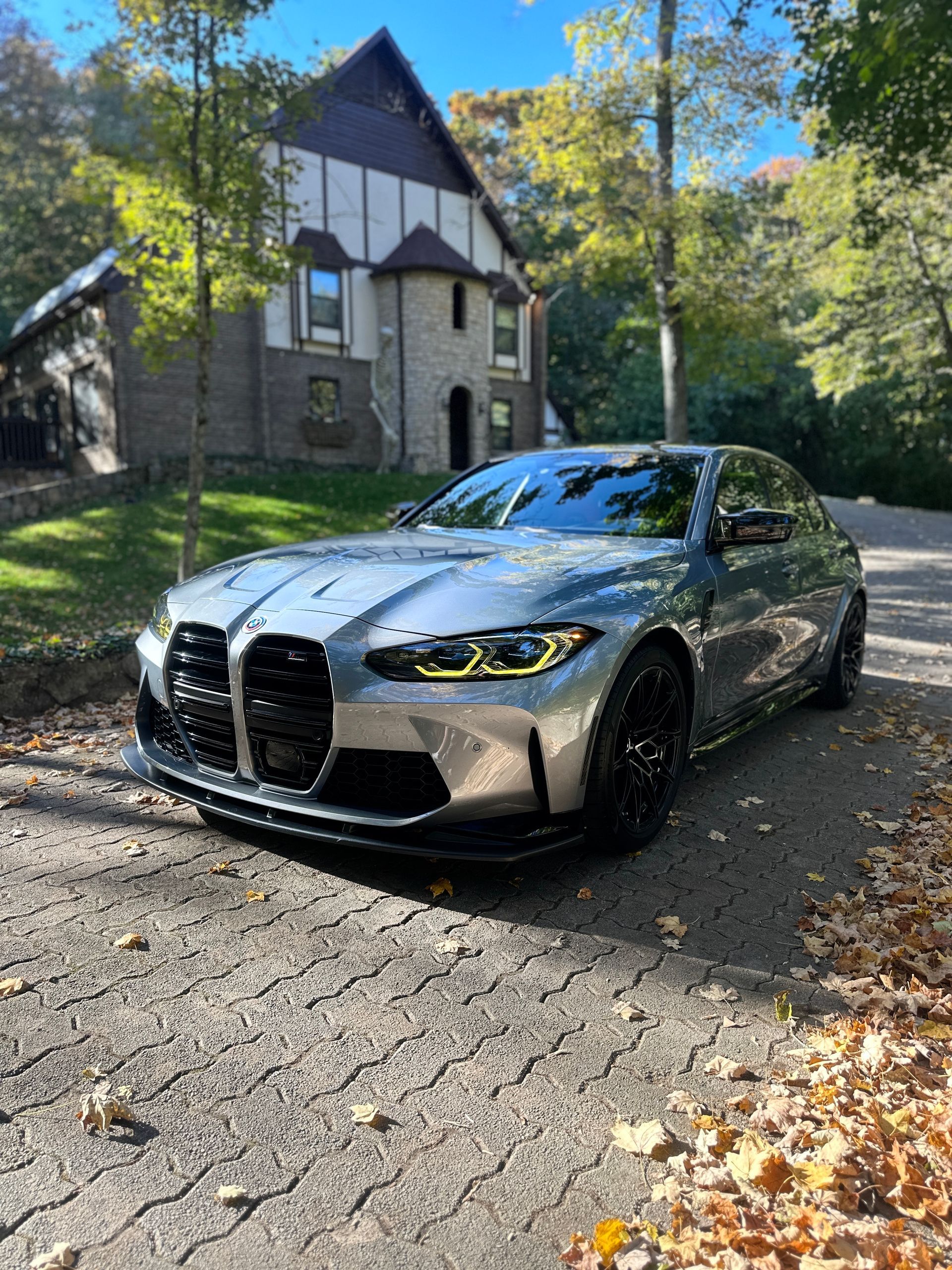 Silver BMW M3 parked on a paved driveway in front of a large stone house with fall foliage surrounding it.