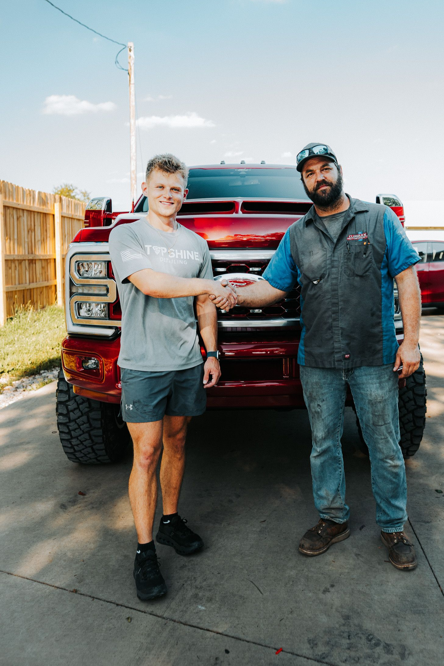 Two men standing in front of a red pickup truck, shaking hands on a sunny driveway.