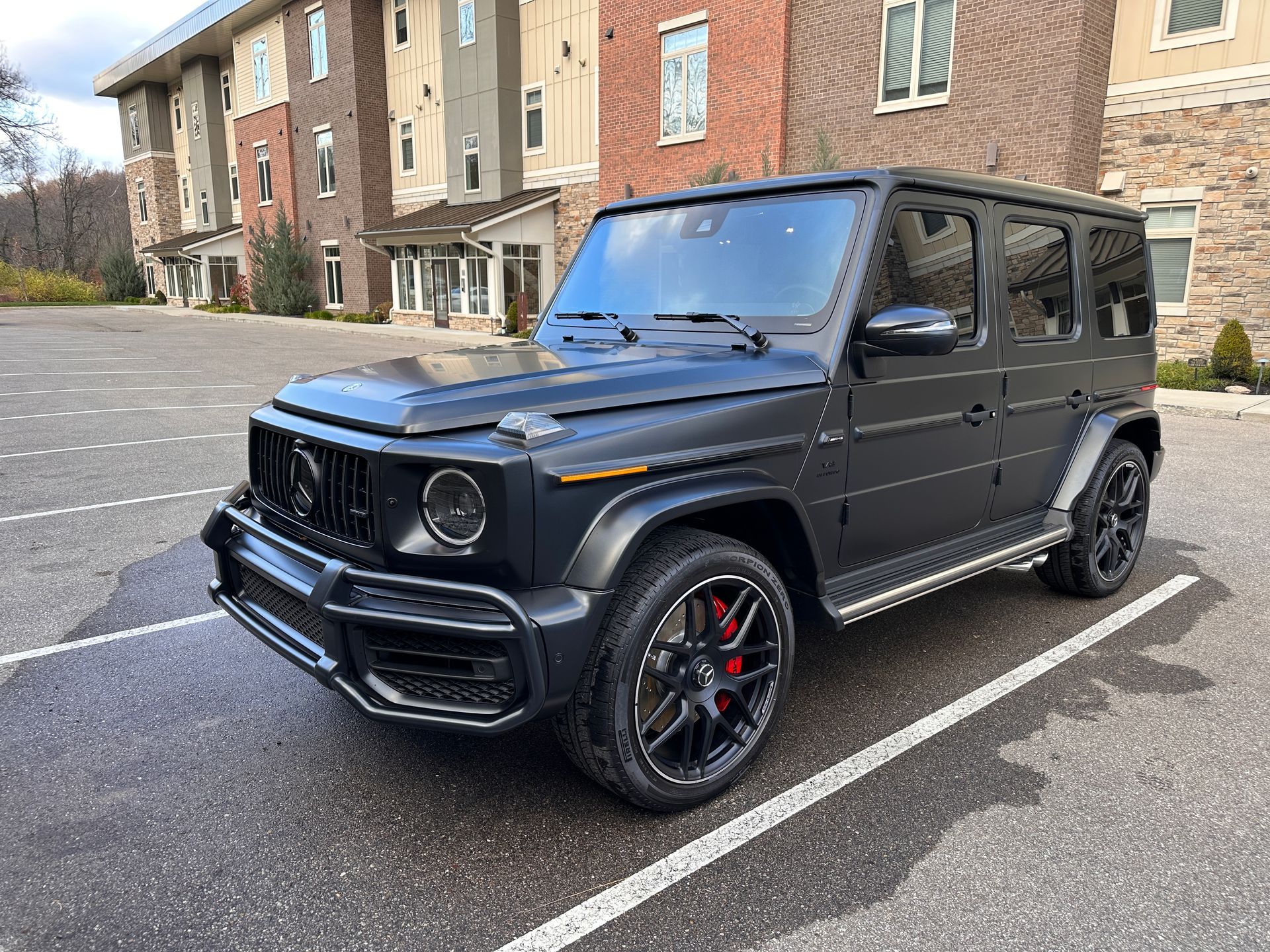 Black Mercedes-Benz G-Class SUV parked on wet asphalt in front of a brick building. The SUV has black rims with red brake calipers.