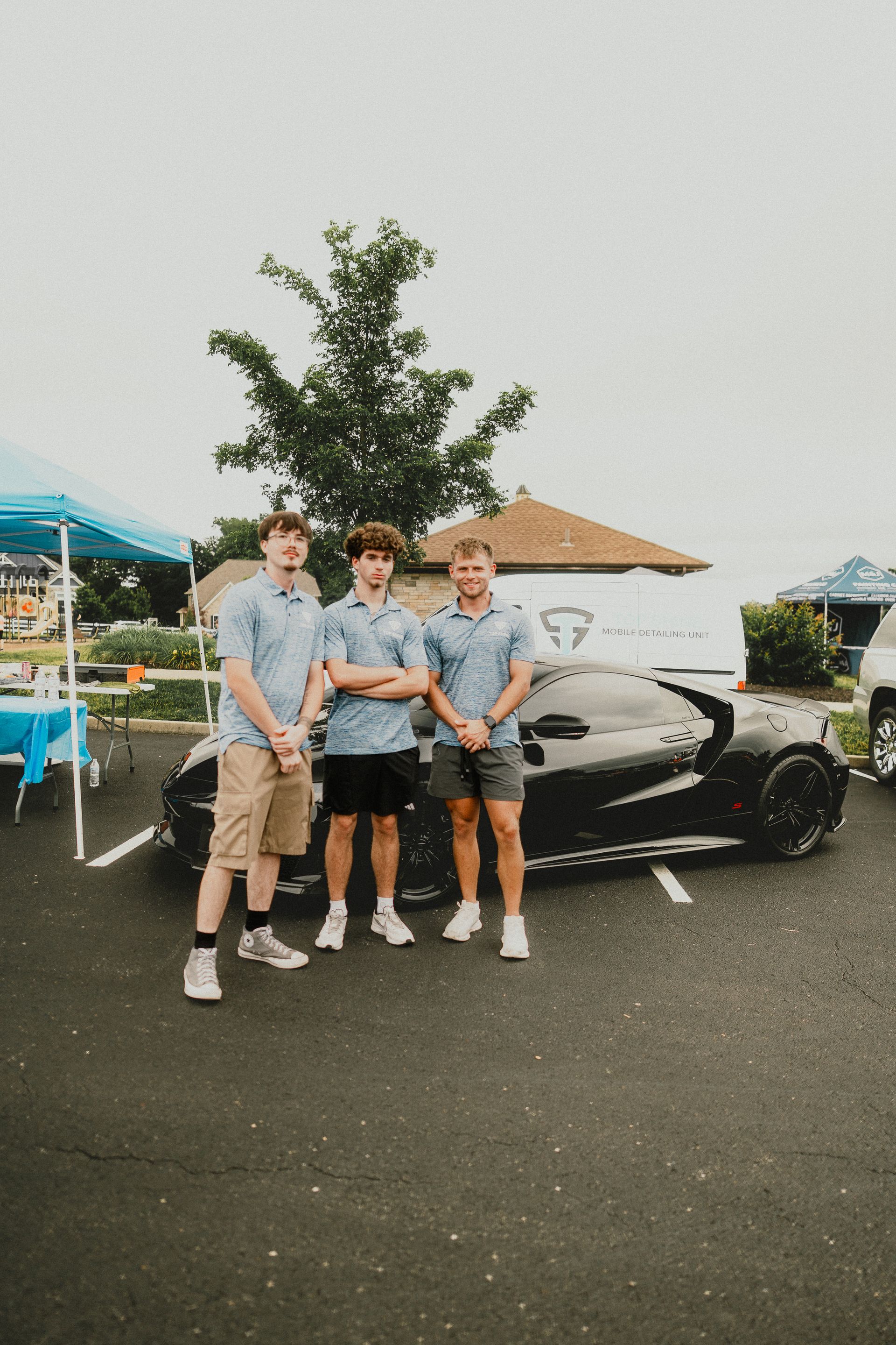 Three people standing in front of a black sports car at an outdoor car meet, holding trophies.