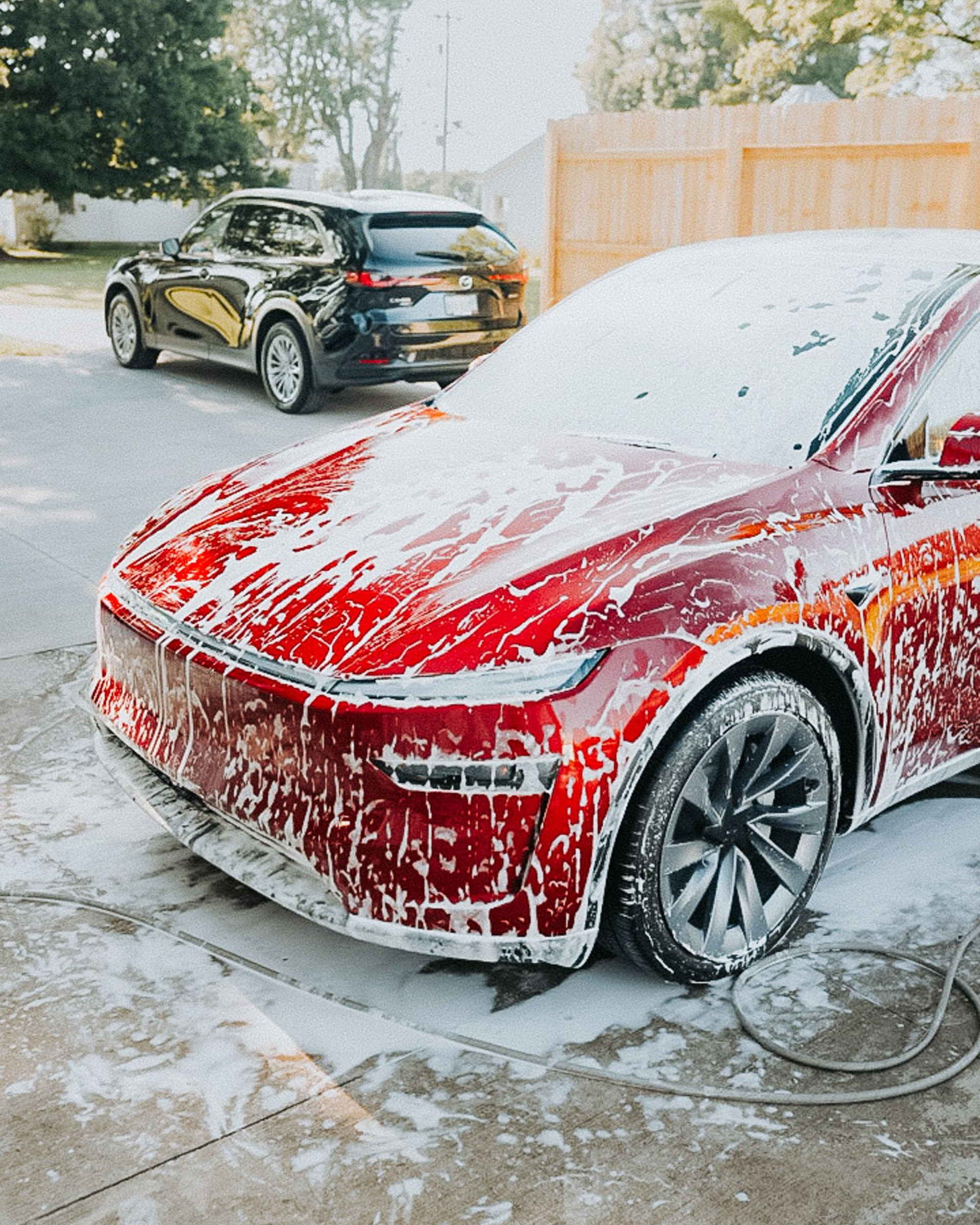 Red car covered in foam being washed outdoors, with a black SUV in the background.