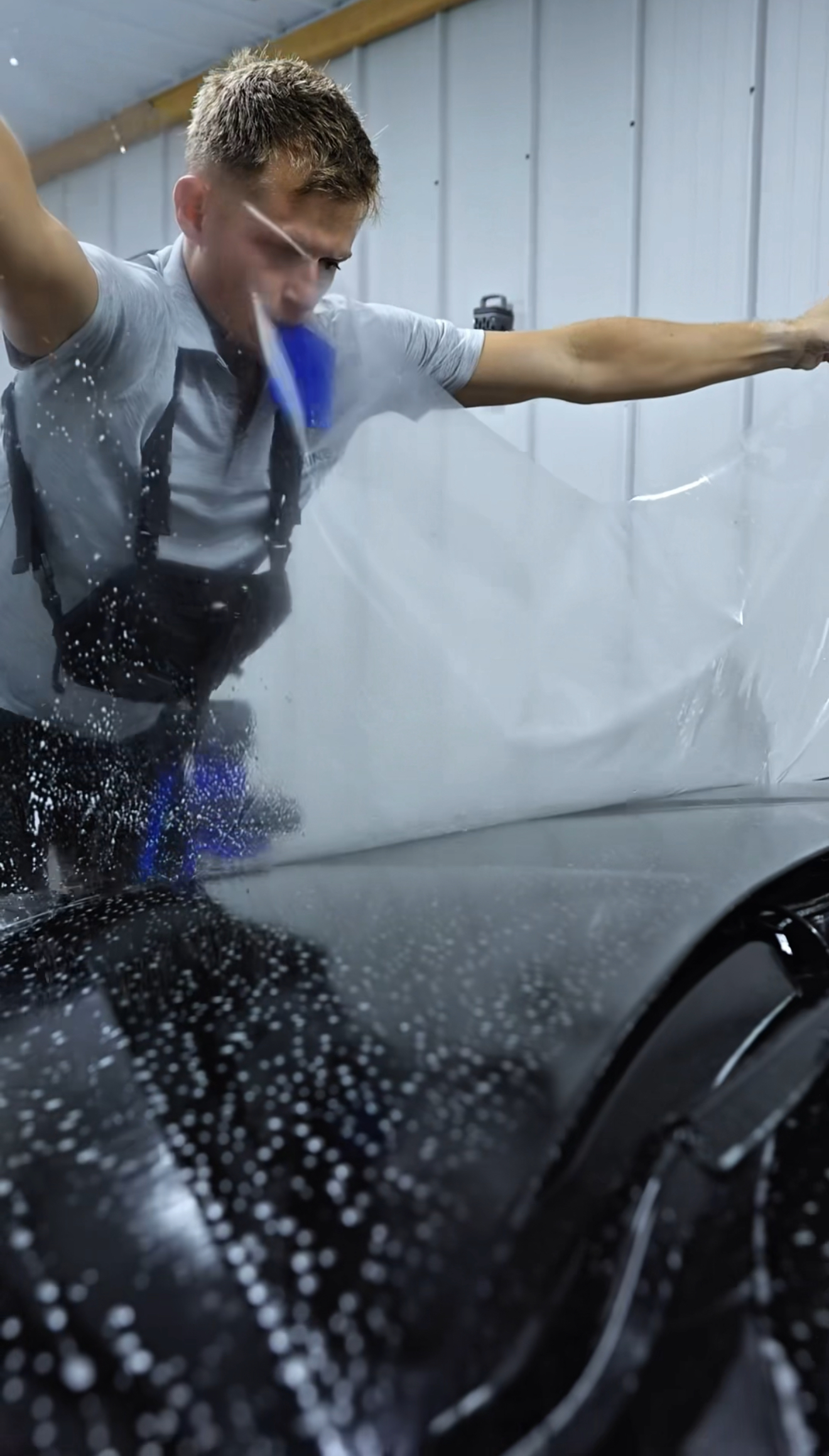 Man polishing a black car in a garage-style detailing bay, wearing a hair net and gray shirt