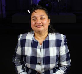 Una persona sonriente de cabello oscuro lleva un blazer a cuadros azul marino y blanco sobre un fondo oscuro y minimalista.