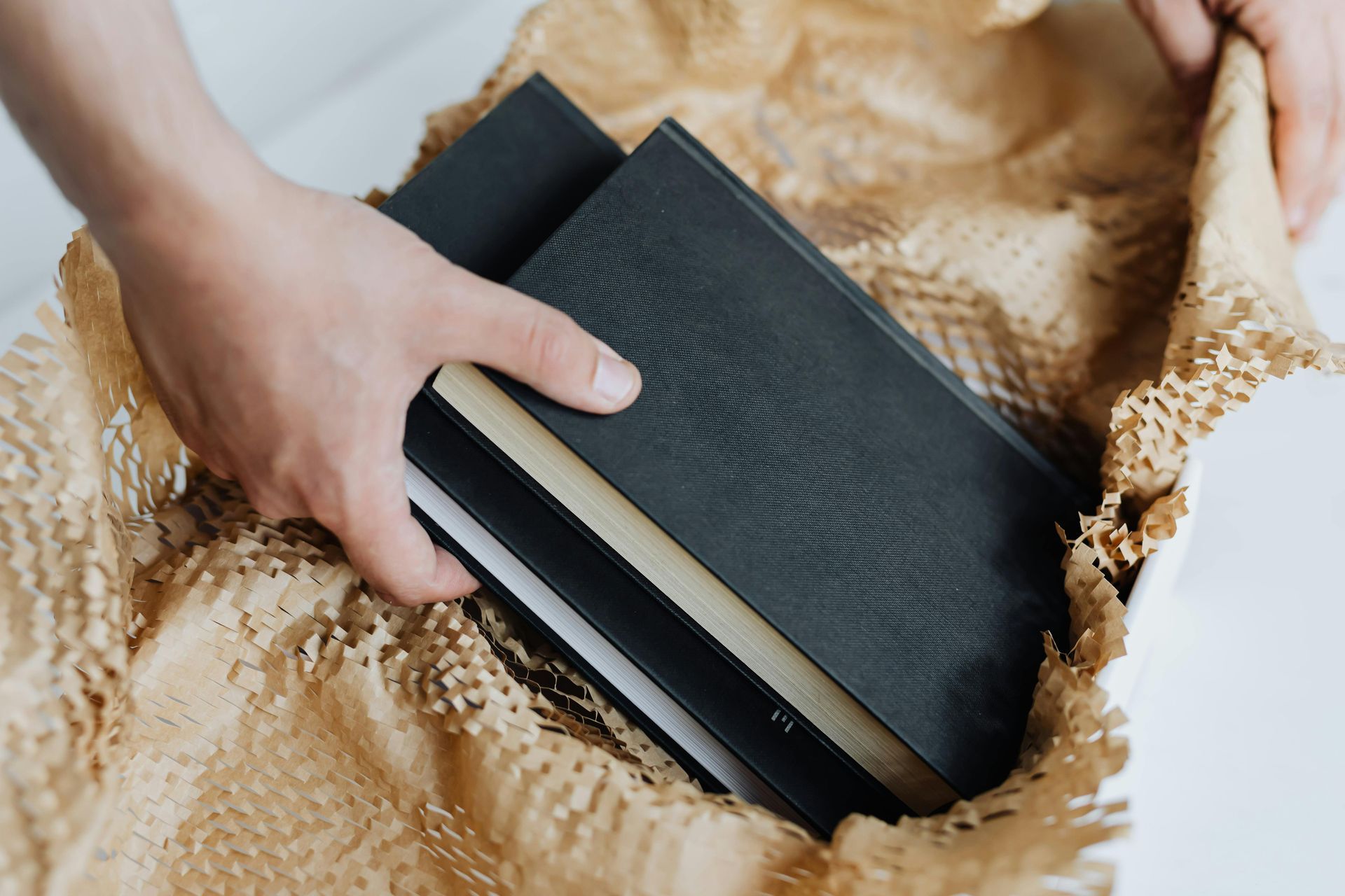 Hands holding black books nestled in brown packing paper.