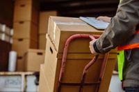 Delivery person pushing a hand truck loaded with boxes toward a truck. Person is holding clipboard.