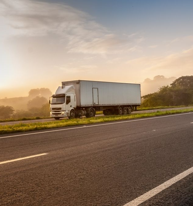 White semi-truck driving on a highway at sunset, with a backdrop of trees and a hazy sky.