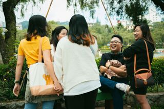 Group of friends chatting and laughing outdoors on a park bench, with trees in the background.