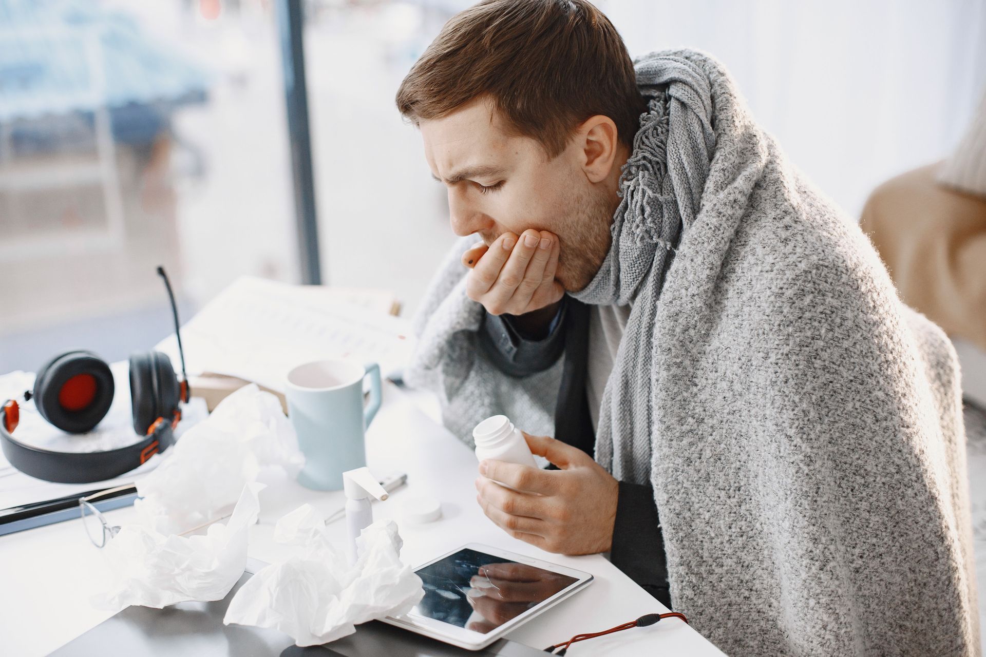 Person wrapped in a gray blanket, blowing their nose beside tissues and a laptop on a desk