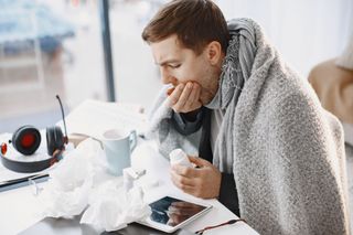 Person wrapped in a gray blanket, blowing their nose beside tissues and a laptop on a desk