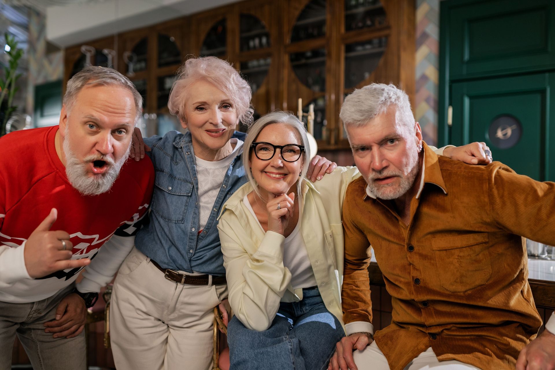 Four smiling friends lean toward the camera in a cozy living room, one holding a red mug.