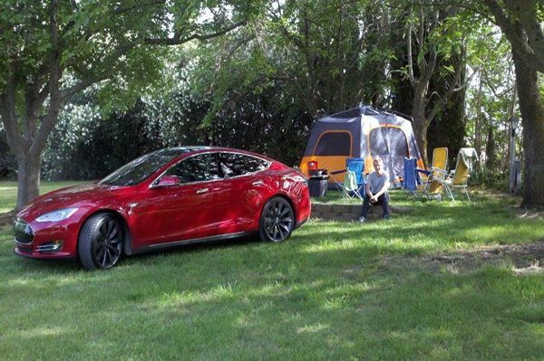 Red Tesla parked next to a campsite with a tent, trees, and person sitting in a chair.