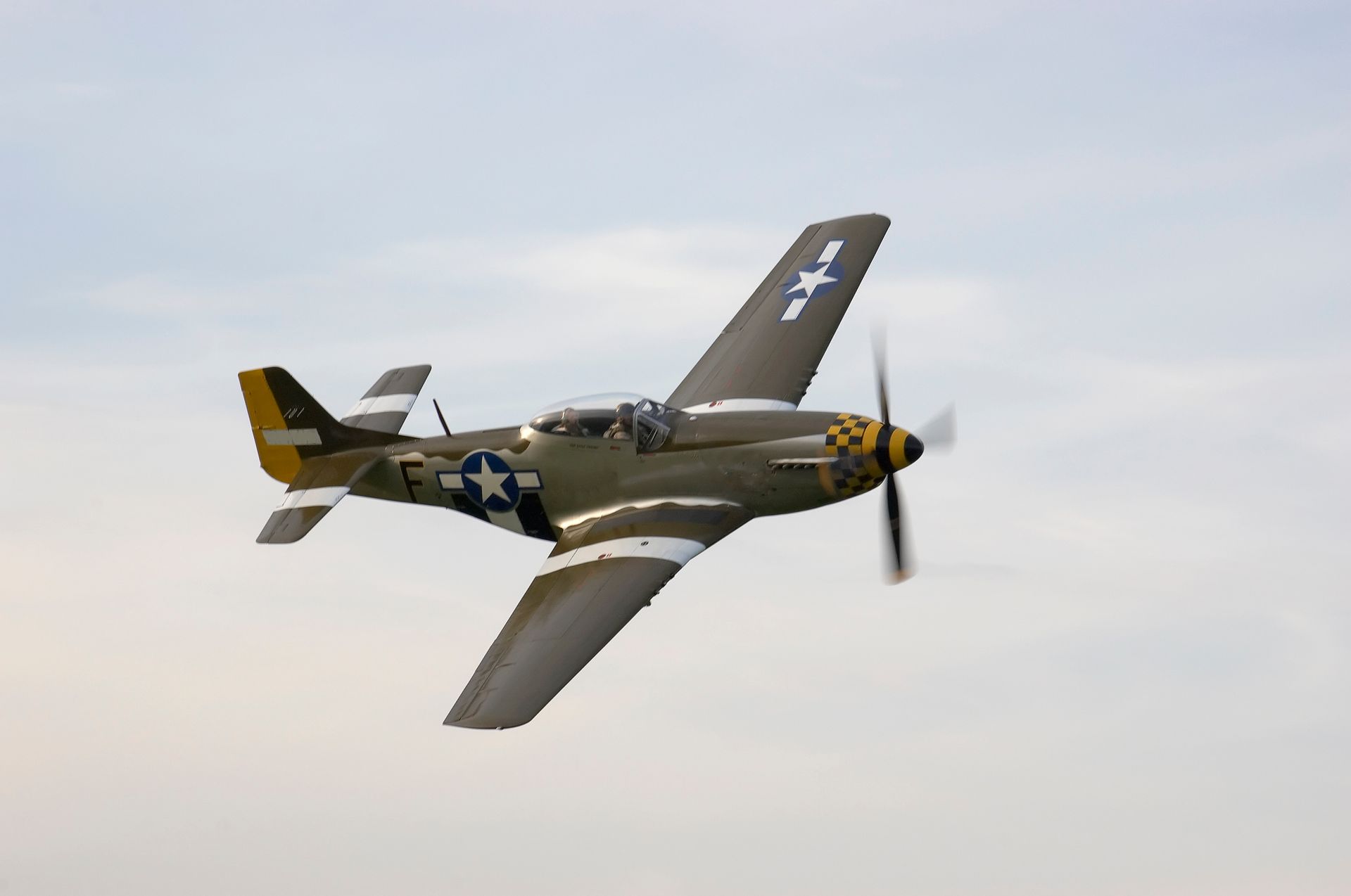 Olive-drab World War II P-51 Mustang fighter plane in flight, white markings, blue sky.