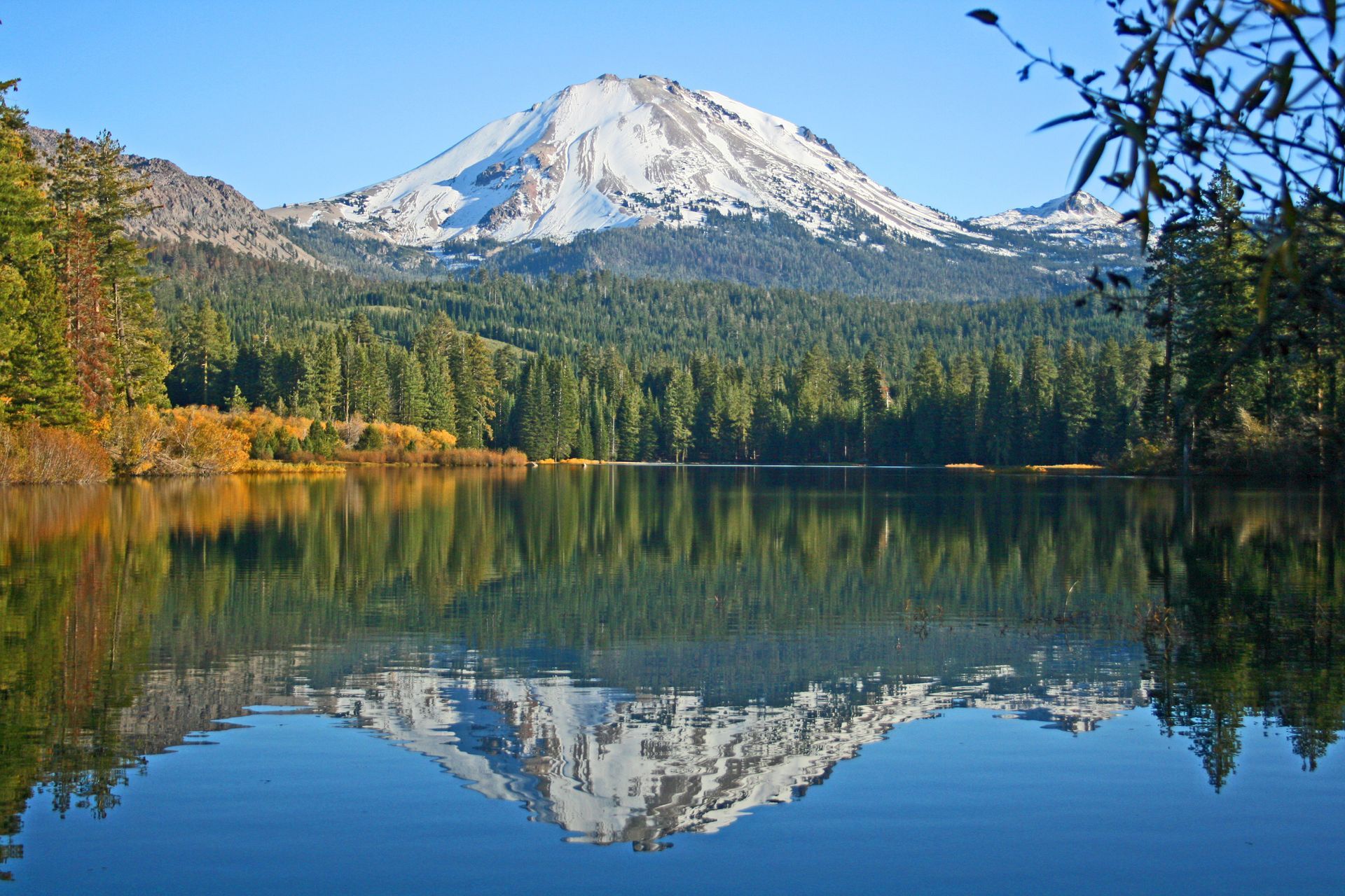 Snow-capped mountain reflecting in a calm lake, surrounded by a forest of green trees under a blue sky.