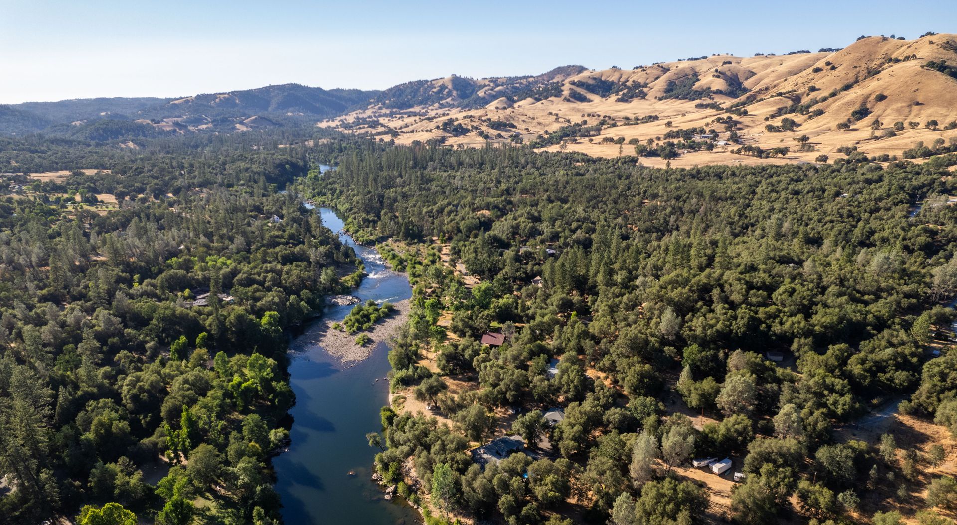 River flows through a forest, with hills in the background under a blue sky.