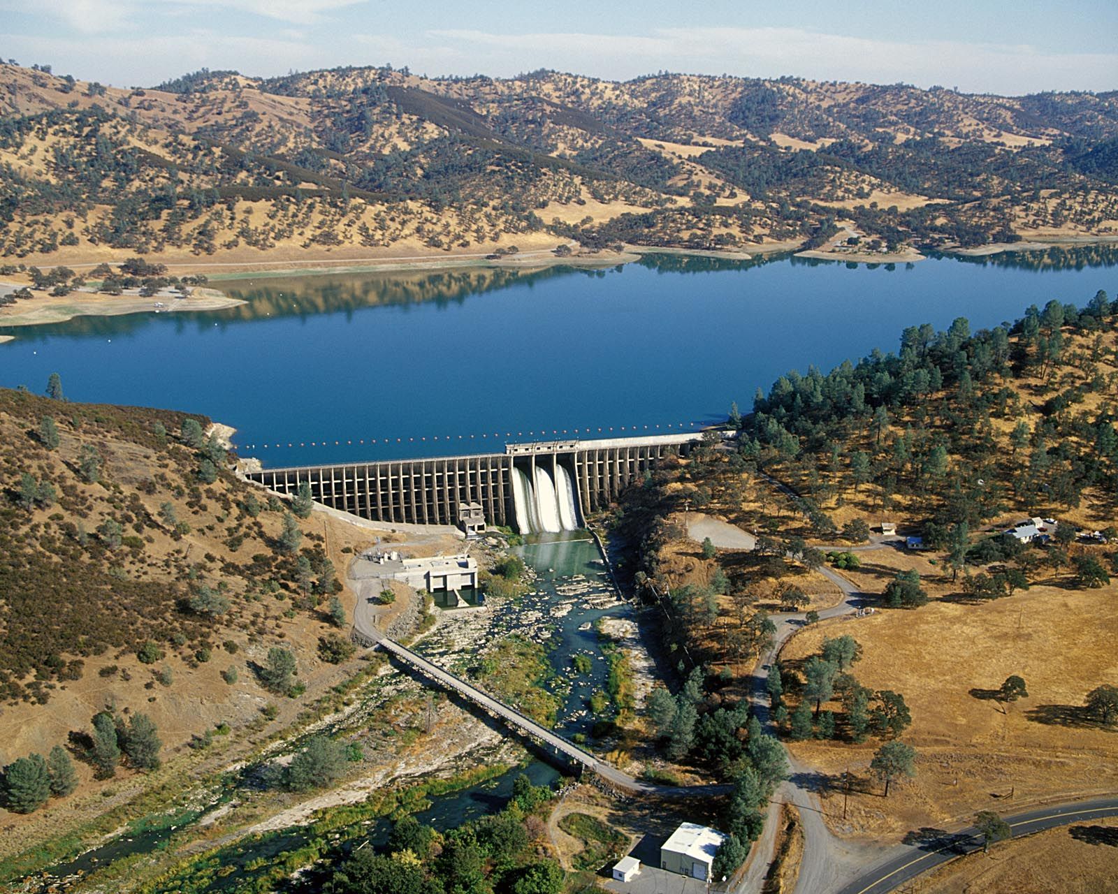 Dam releases water, creating a river. Lake and hills visible. Sunny day.
