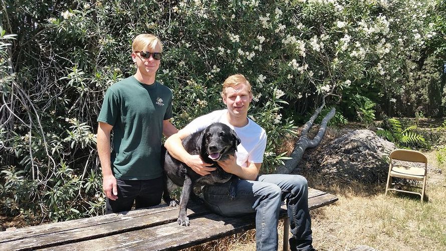 Two young men with a black dog outdoors. One man sits, holding the dog, the other stands. Green foliage in the background.