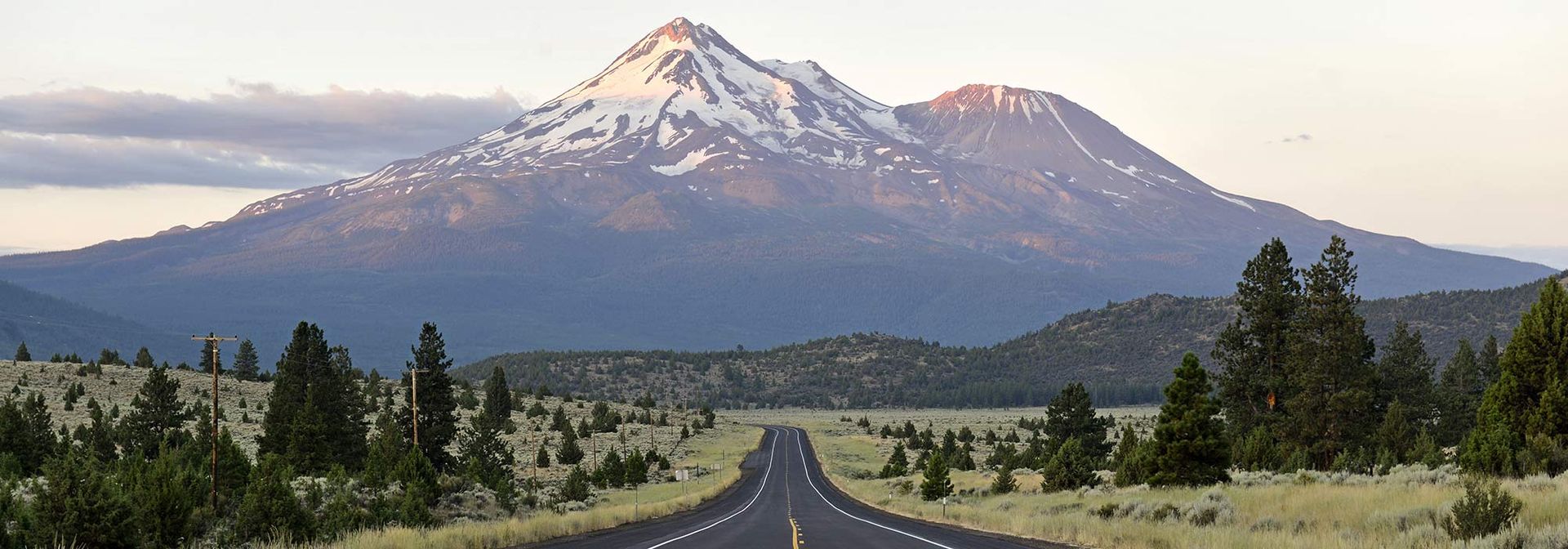 Asphalt road through a grassy field towards snow-capped mountains. Sparse trees line the sides. Sky is hazy.