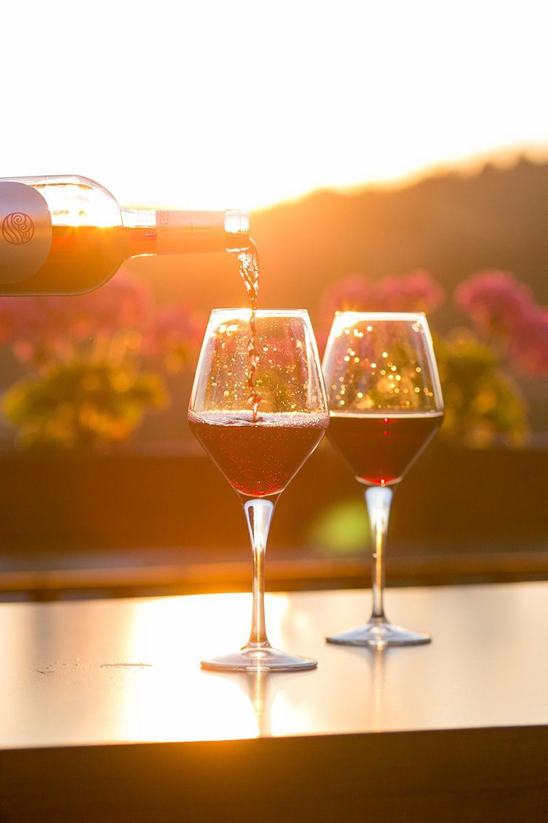 Two wine glasses filled with red wine, being poured from a bottle, sunset background.