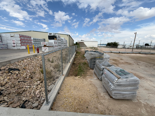 Construction site with stacked materials, gravel path, and chain-link fence under a cloudy sky