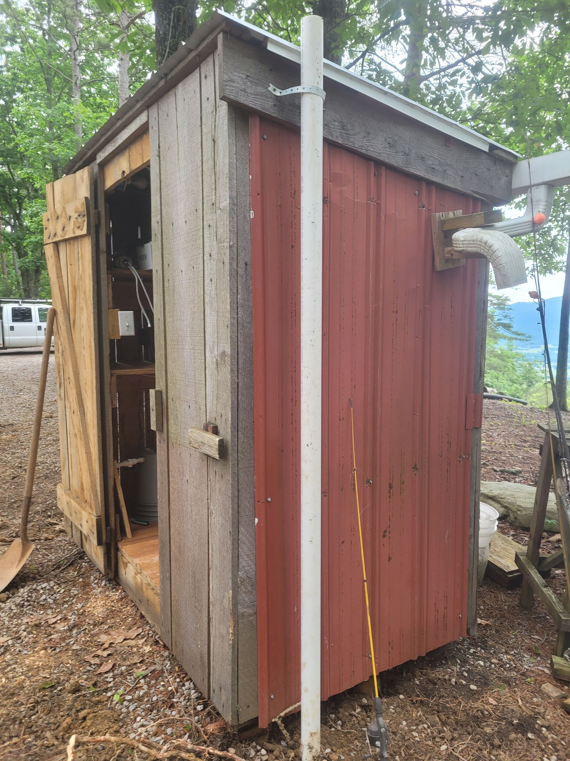 Wooden outhouse with red siding, white pipe, and open door on a wooded hillside.