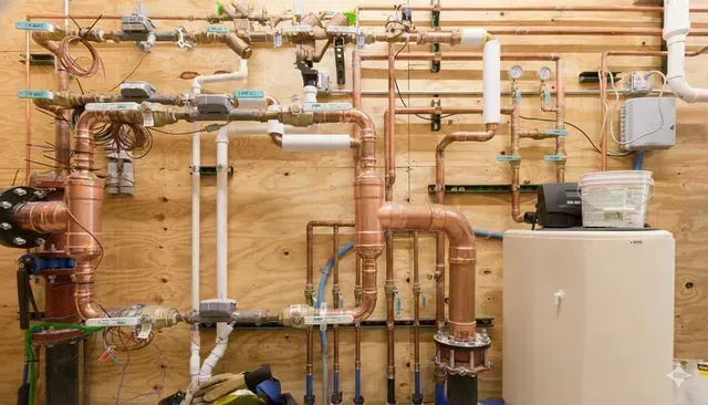 Complex copper and white pipe plumbing system mounted on a wooden wall next to a beige water tank.