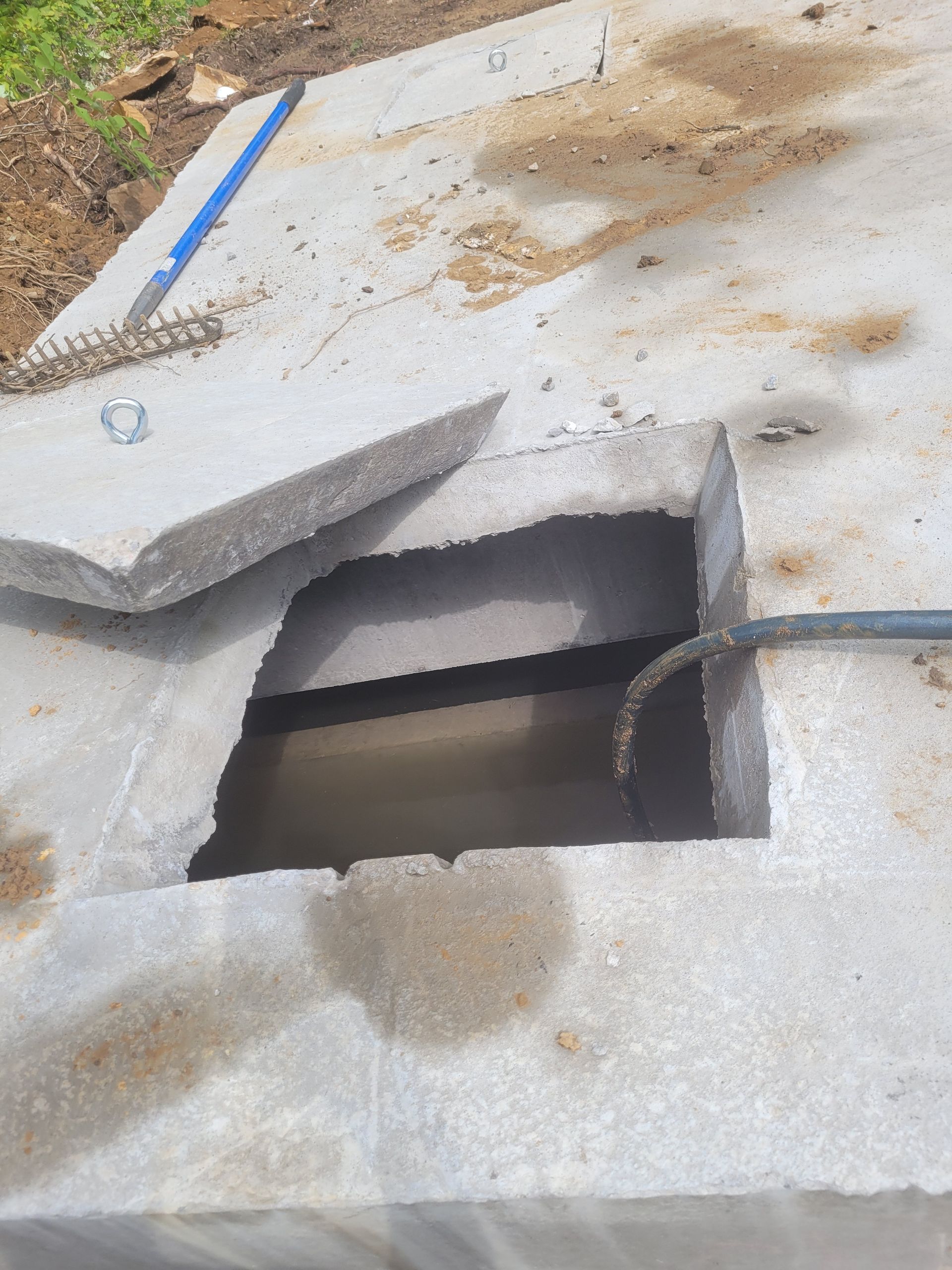 Concrete slab with a rectangular opening, water visible inside. A piece of slab is broken off. A hose is inserted.