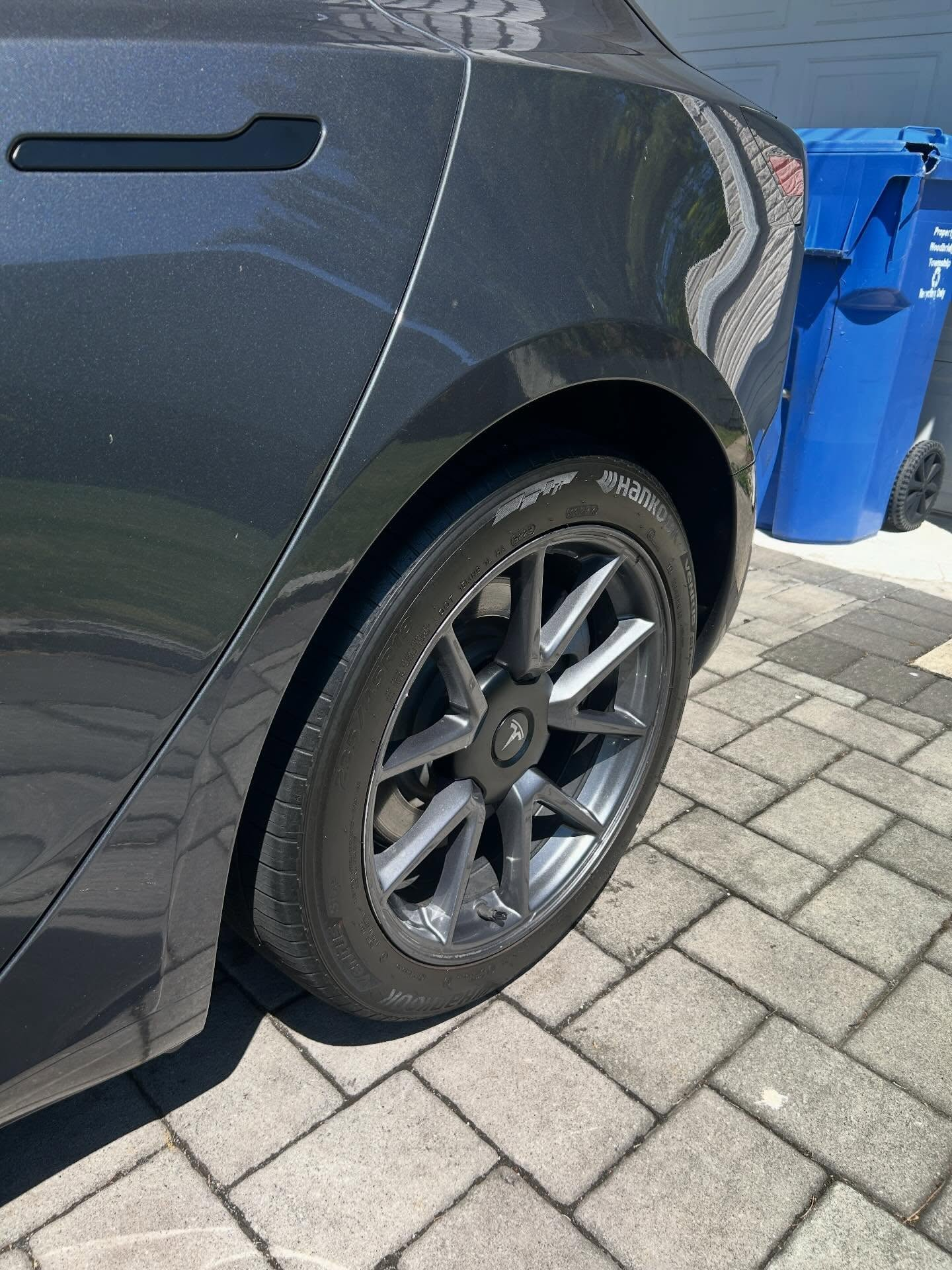 Rear side of a dark gray car parked on a brick driveway beside blue recycling bins.