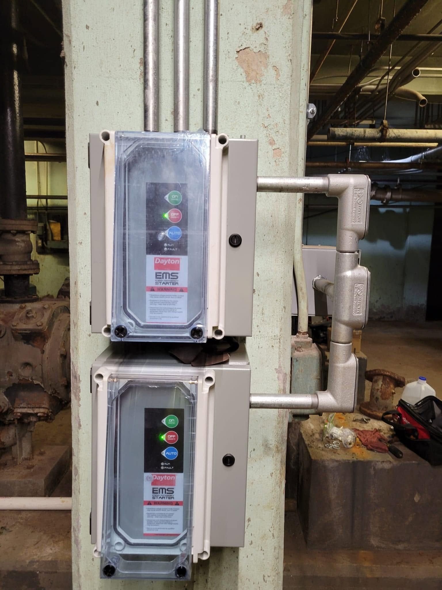 Two wall-mounted industrial control boxes with clear covers on a factory column and pipes.