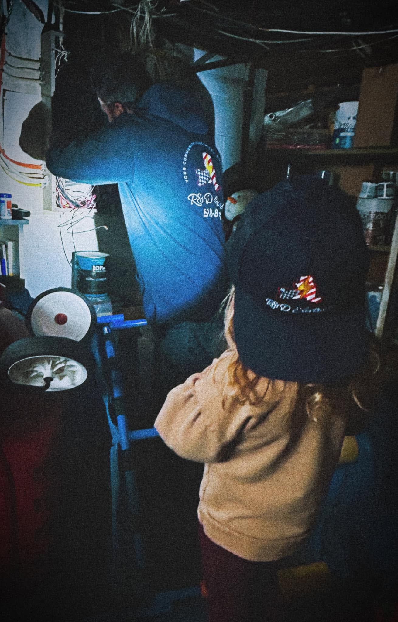 Two people in winter hats and jackets stand in a dim basement near shelves and pipes.