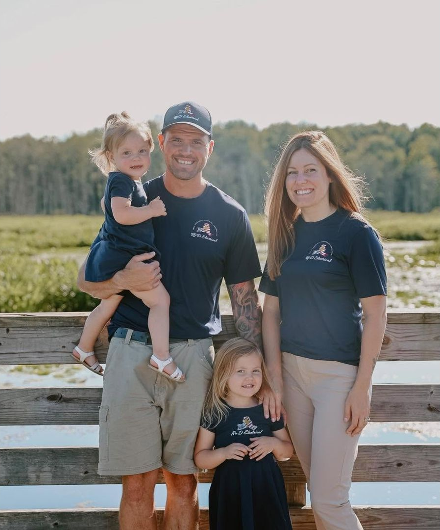 Family of four standing outdoors by a wooden fence, wearing matching navy shirts, smiling at the camera.