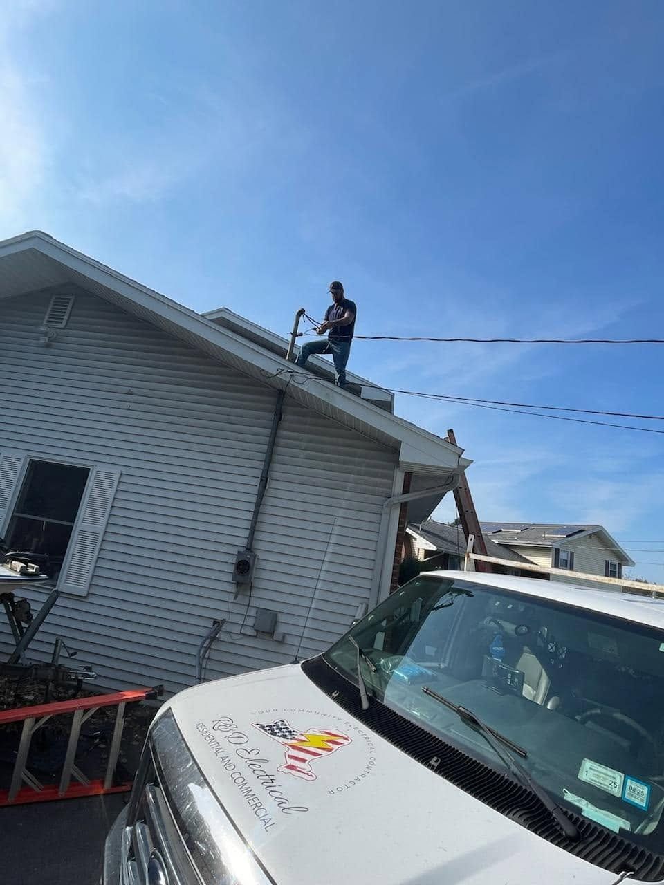 Worker on a roof above a white house, with a white vehicle in the foreground after storm damage.