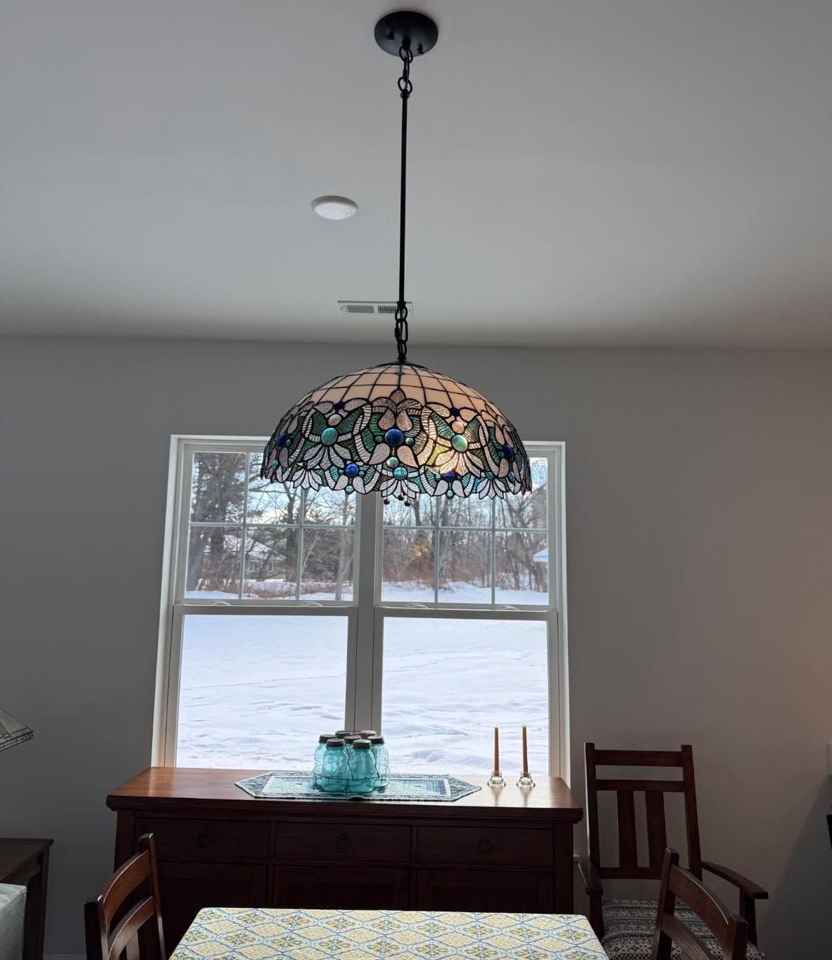 Dining room with dark wood table, stained-glass pendant light, and snowy view through large window.
