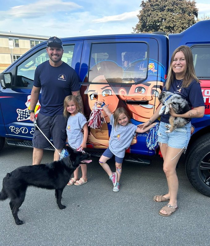 Family posing with a dog beside a blue vehicle with a cartoon character wrap.