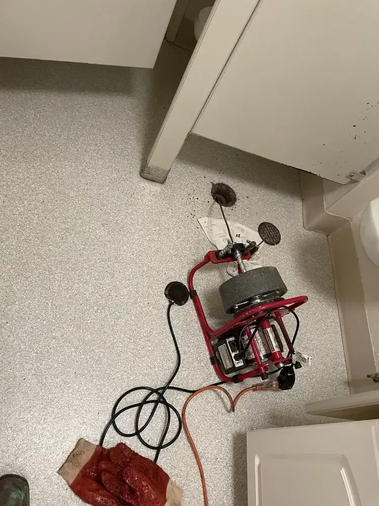 Red wheeled robot on a floor near cabinets, with wires attached and a red cloth beside it