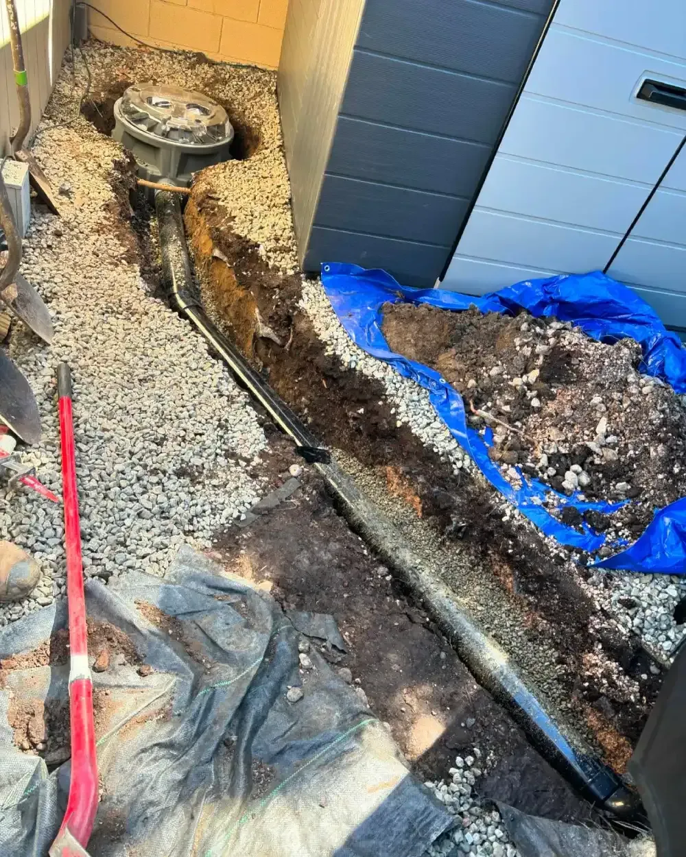 Outdoor trench beside a building, with gravel, blue tarp, and a long black pipe laid in the ground.