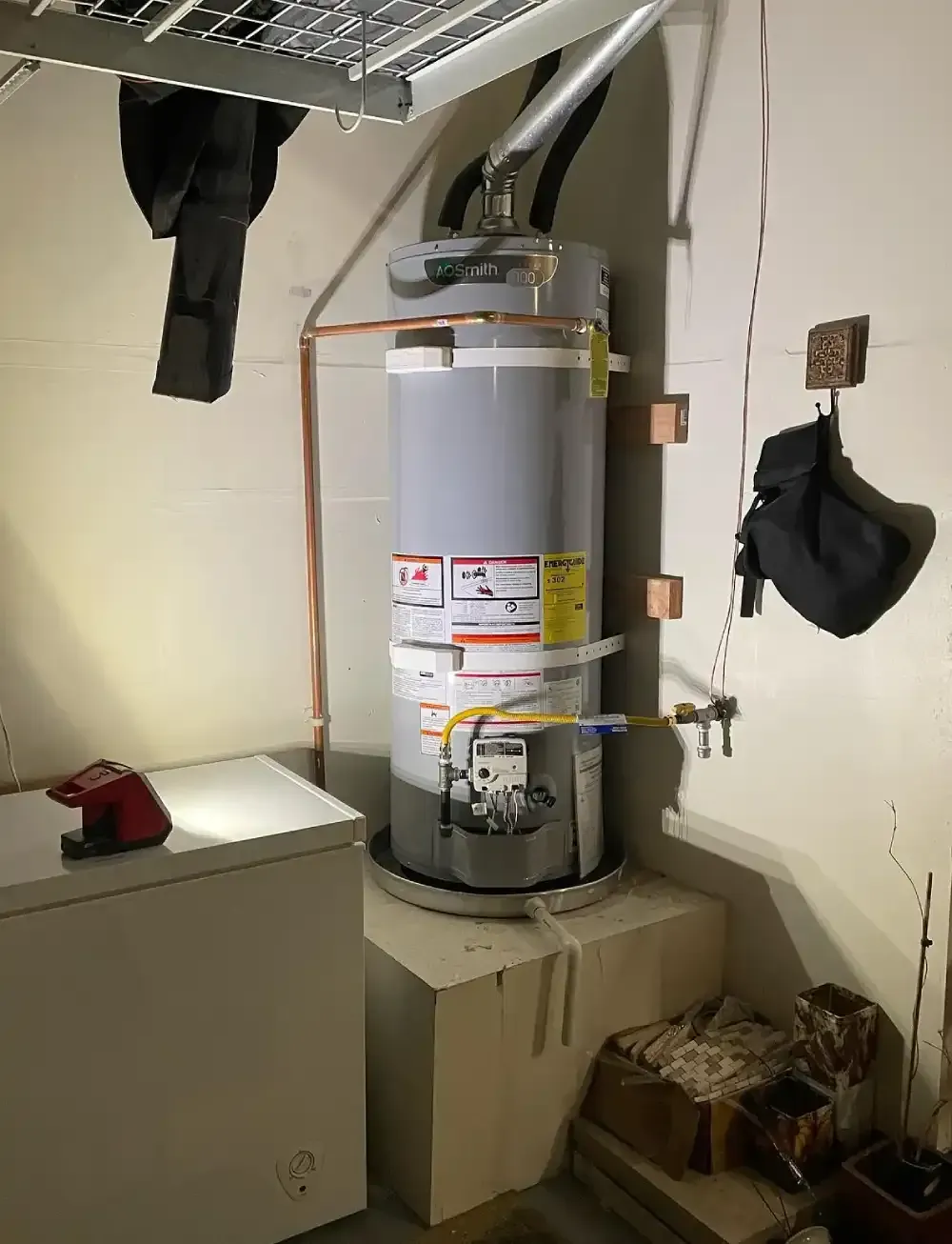 Water heater in a utility closet with nearby pipes, cabinets, and stored items.