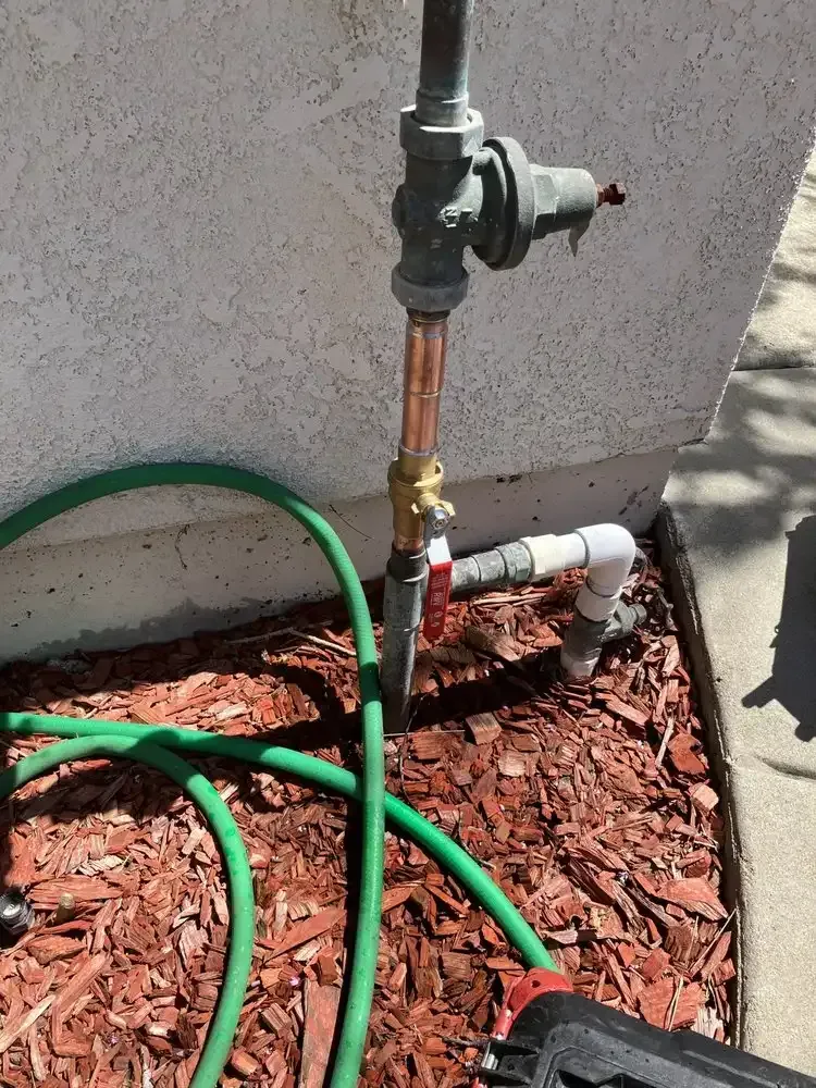 Outdoor hose bib with green garden hose connected to a copper pipe against a wall, over red mulch.
