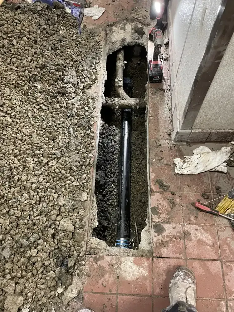 Open floor trench exposing black plumbing pipes beside a tiled floor and gravel in a construction area