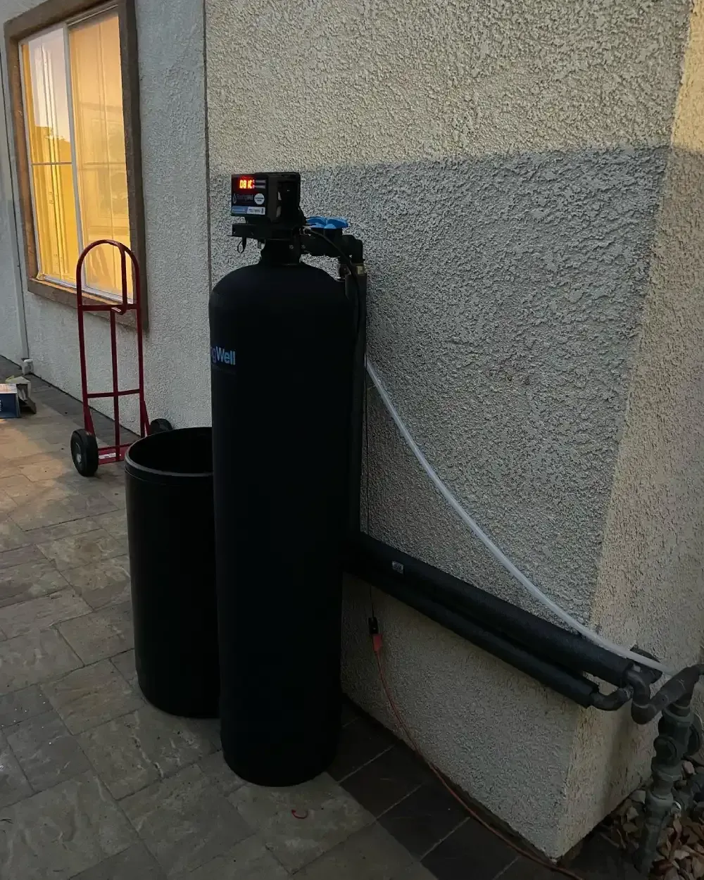 Two black water softener tanks beside an exterior wall, with a red dolly nearby.