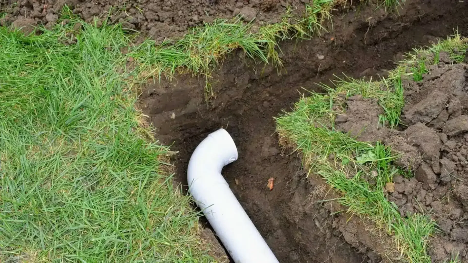 White drain pipe protruding from a dirt trench beside green grass.