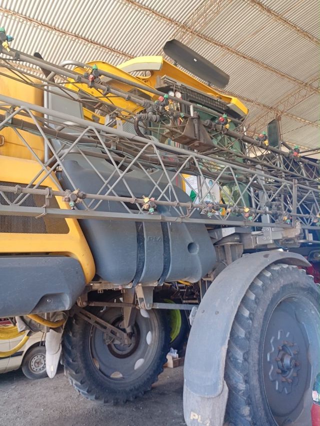 Pulverizador agrícola amarillo montado en un camión grande dentro de un cobertizo, con los brazos de pulverización plegados.