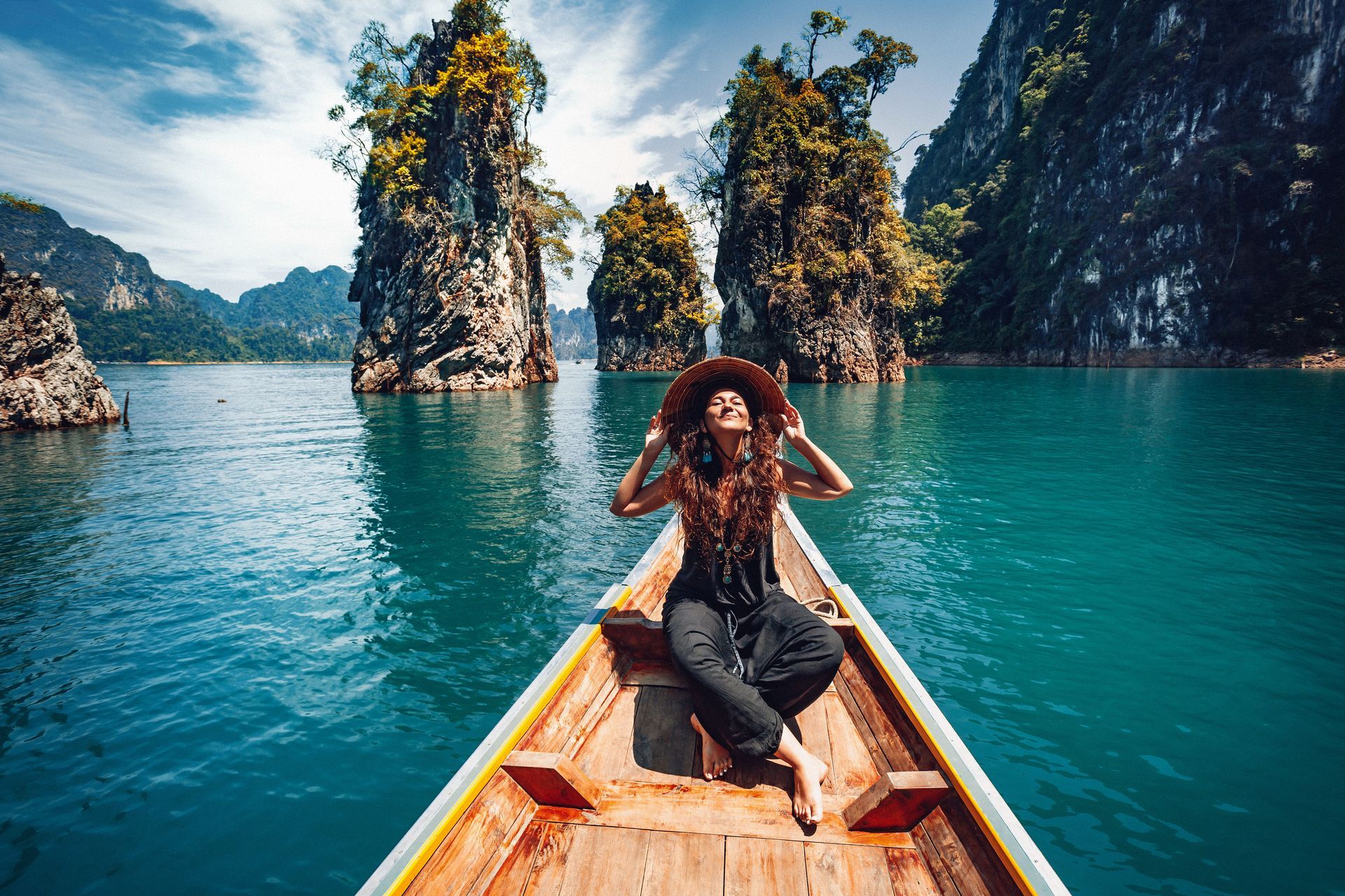 Person on a wooden boat in turquoise water, with tall rocky islands and a forested cliff.