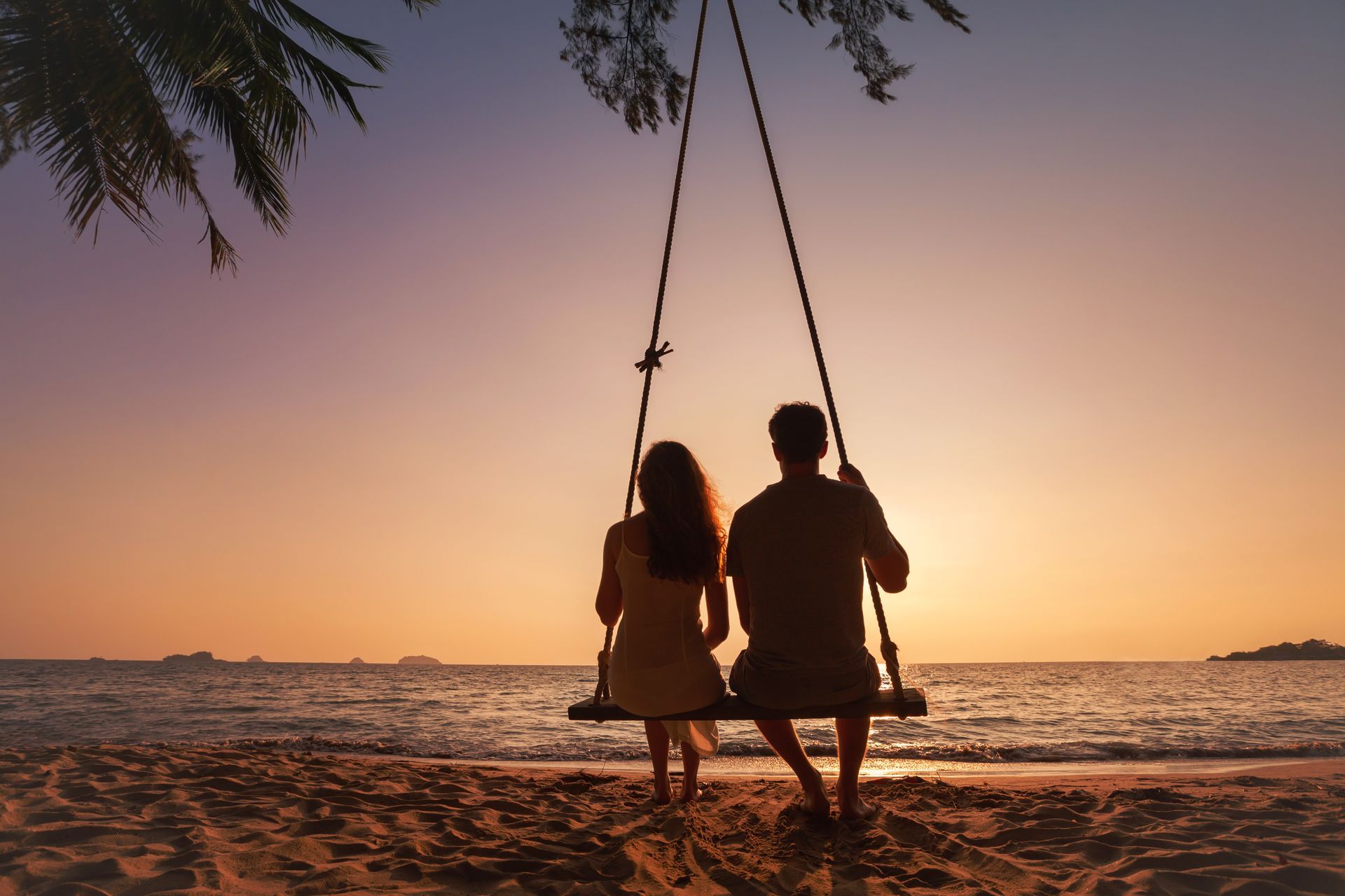 Two people on a swing at sunset by the beach, with palm leaves overhead and calm water behind.