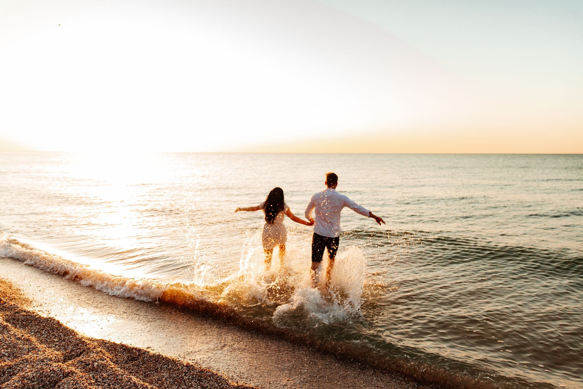 Two people run into the ocean at sunset, splashing through shallow water on a pebbled beach.