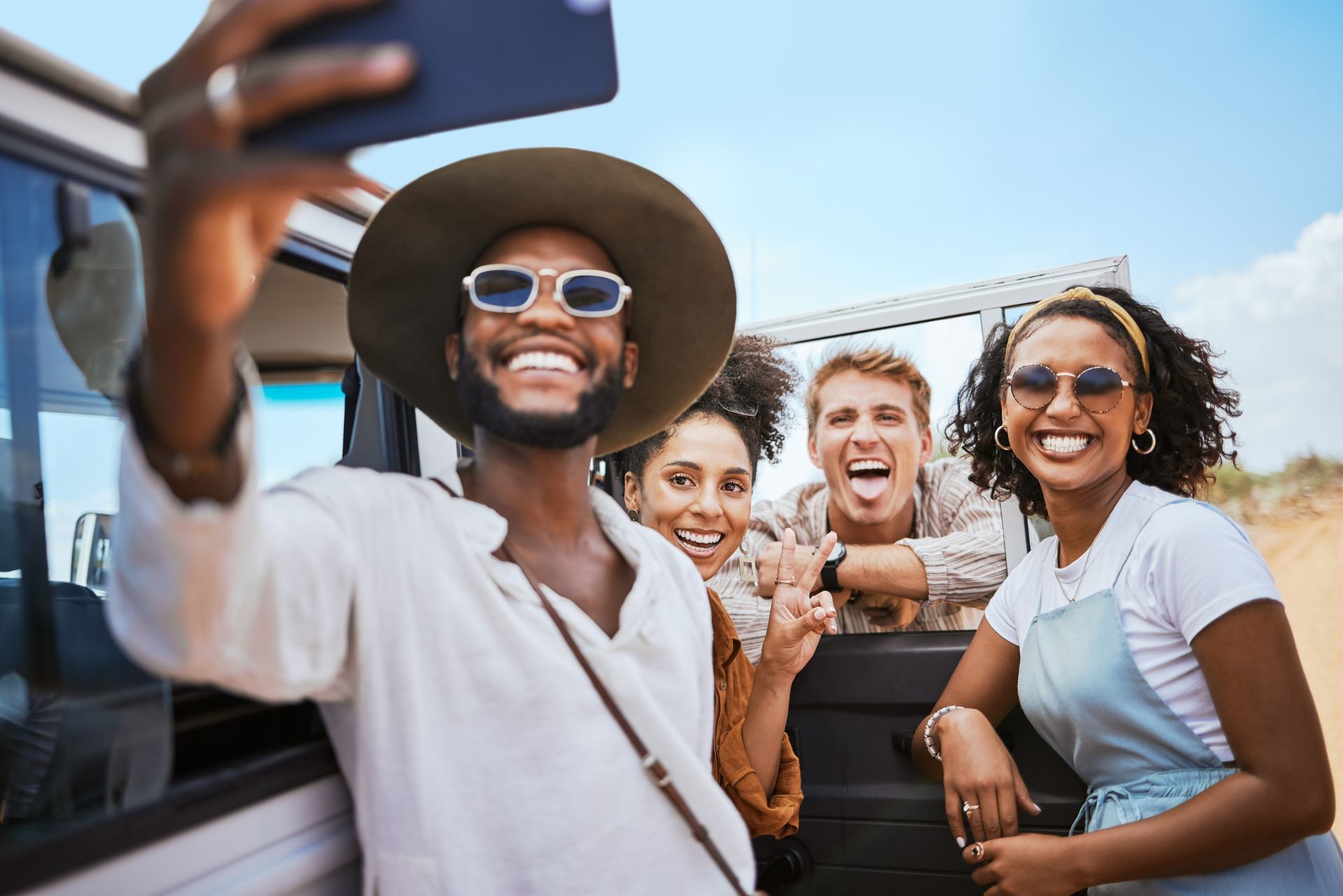 Four friends smiling on a road trip, taking a selfie near a vehicle under a bright blue sky.