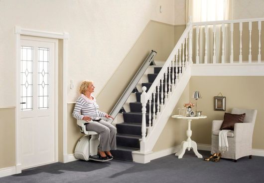 Donna che utilizza un montascale su una scala con pianerottolo; pareti beige, porta bianca, tappeto, sedia.