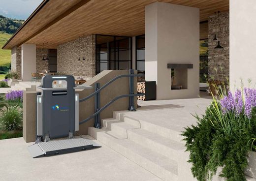 Elevatore per sedie a rotelle grigio sui gradini esterni, accanto a una casa con muri in pietra e soffitto in legno.