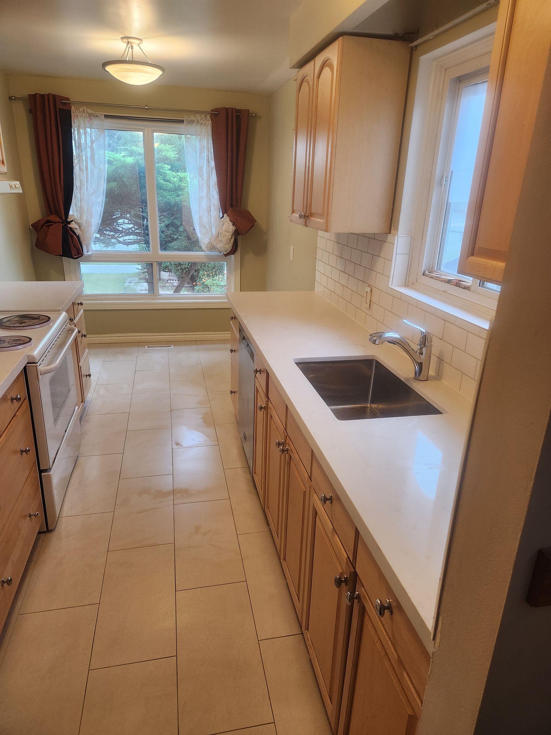 Kitchen with light wood cabinets, white countertops, and a window with curtains.