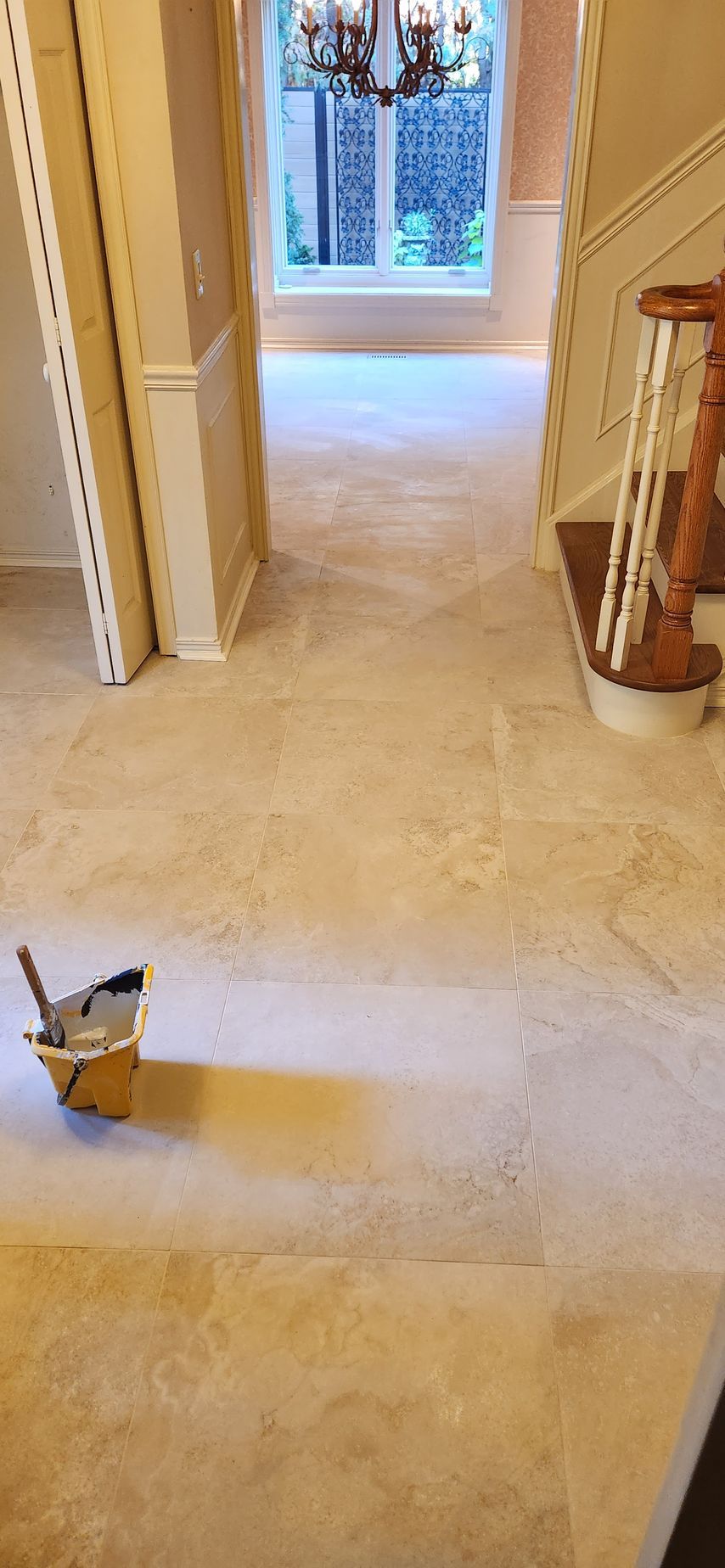 A room with beige tile floors, a window at the end, and a staircase to the right. A bucket of tools is on the left.