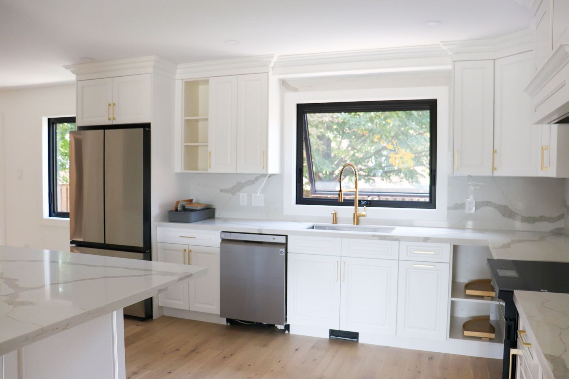 Modern white kitchen with stainless steel appliances, marble countertops, and a black-framed window.